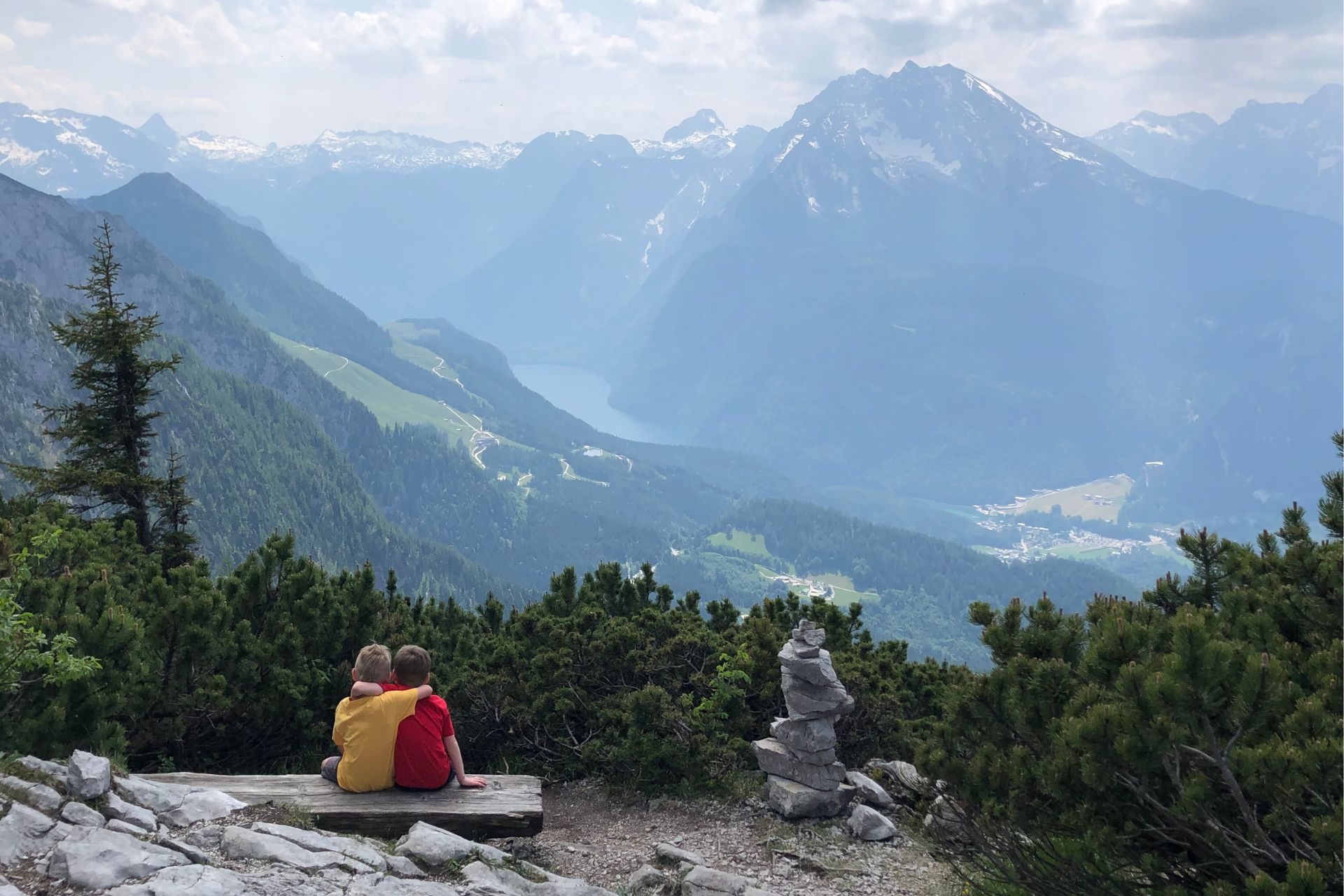 Two children, one in a yellow shirt and the other in red, sit on a wooden bench embracing as they overlook a breathtaking view of the Bavarian Alps near the Eagle's Nest on the Germany-Austria border. The scene features rolling green hills, winding roads, and a serene mountain lake nestled among towering, snow-capped peaks under a hazy sky.