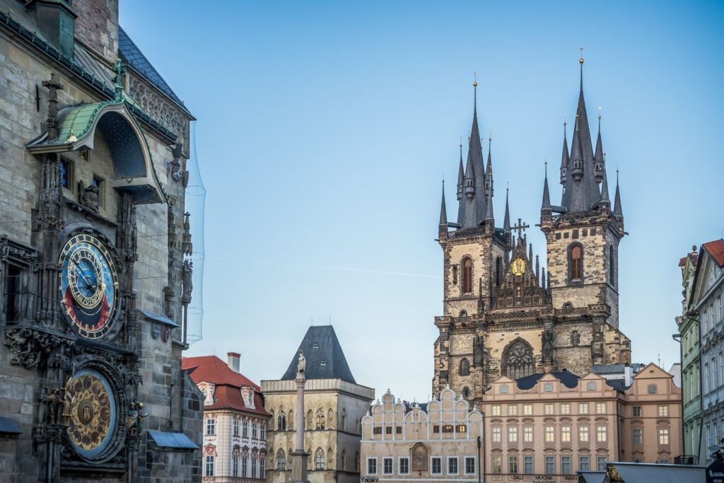 View of Prague’s Old Town Square featuring the Astronomical Clock on the left and the Gothic spires of the Church of Our Lady before Týn in the background. Colorful historic buildings with ornate facades line the square under a clear blue sky.