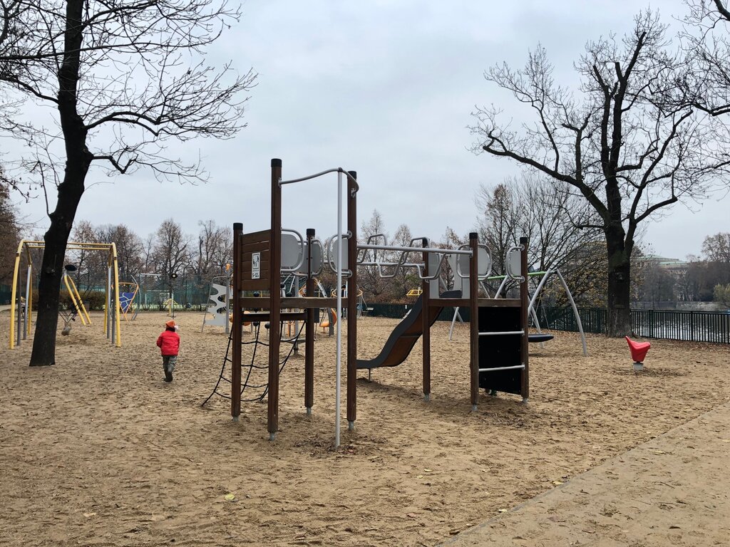 A nearly empty sand-covered playground in Prague features a climbing structure and swings, with a child in a red coat walking alone among bare trees.