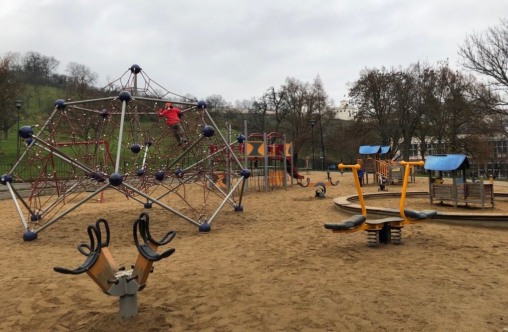 A large sandy playground features a geometric rope climbing structure, colorful play equipment, and a seesaw with spring-mounted seats. A child in a red jacket is seen climbing high on the web-like ropes under an overcast sky.