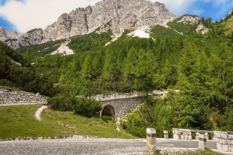 A scenic stone bridge along the Vršič Pass, surrounded by lush greenery and towering mountain peaks in the Julian Alps. The winding cobblestone road curves gently in the foreground, leading towards the bridge and the rugged cliffs beyond. The landscape is a mix of evergreen trees and alpine meadows, with patches of snow clinging to the rocky slopes.