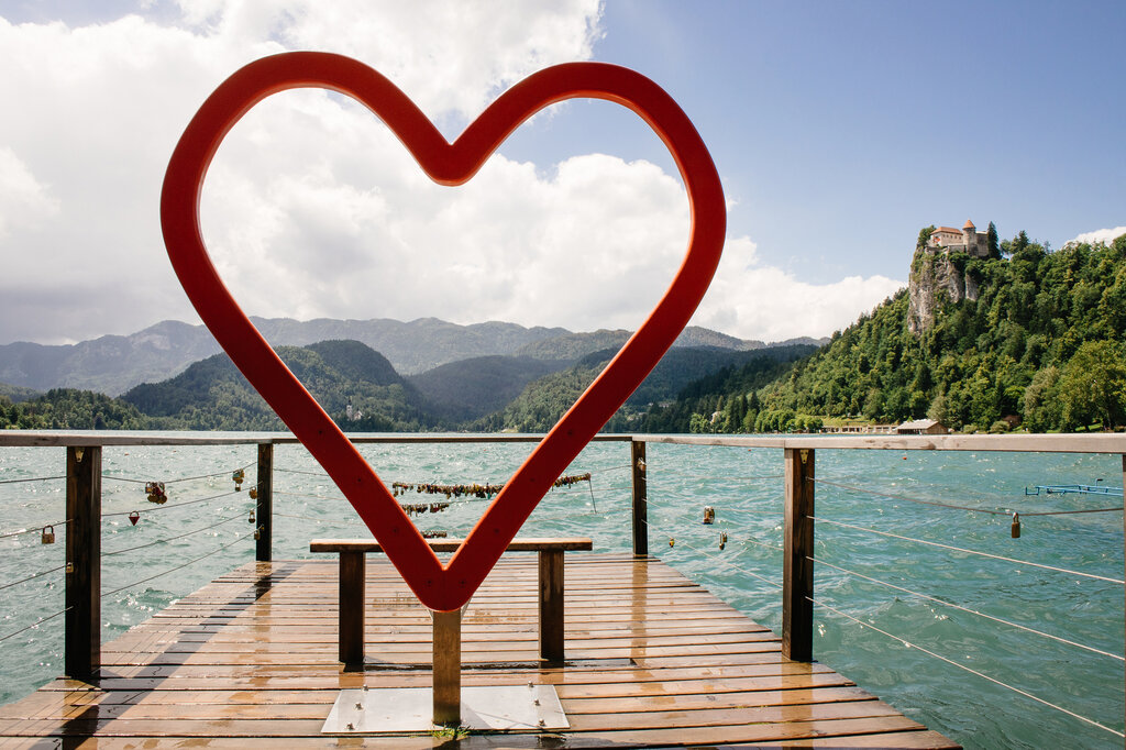 A large red heart-shaped frame stands on a wooden pier overlooking the turquoise waters of Lake Bled. Love locks are attached to the pier’s railing, symbolizing romance. In the background, Bled Castle sits atop a rocky hill, surrounded by lush greenery, with mountains and a partly cloudy sky adding to the scenic view.