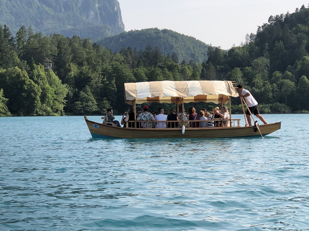 A traditional Pletna boat glides across the turquoise waters of Lake Bled, carrying a group of passengers under a beige-striped canopy. A standing boatman, known as a "Pletnar", skillfully rows the boat using a long wooden oar. Lush green forests and mountains form a scenic backdrop, enhancing the tranquil atmosphere of the lake.