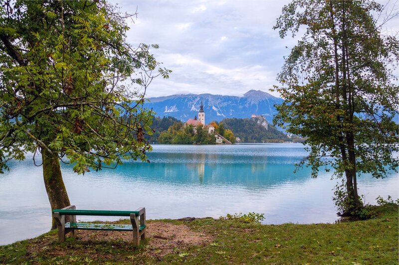 A peaceful lakeside scene at Lake Bled, with a wooden bench under a tree framing the view of Bled Island in the distance. The island, home to the iconic Church of the Assumption, sits in the middle of the lake, surrounded by calm, turquoise waters. In the background, majestic mountains rise beneath a cloudy sky, adding to the tranquil and picturesque atmosphere.