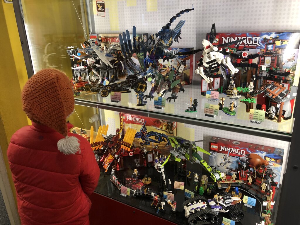 a child in a red coat and orange santa hat looks at a display of ninjago lego on a shelf