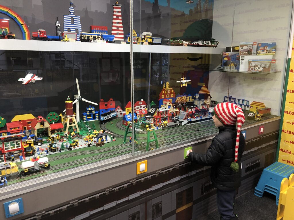A child in a red-and-white striped hat looks at an elaborate LEGO city display behind glass, featuring a miniature town with buildings, vehicles, a train, and a wind turbine. The scene is full of color and tiny details, creating a lively toy world.