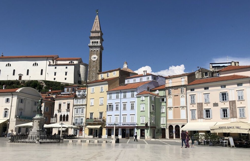 Tartini Square in Piran, Slovenia, on a sunny day, surrounded by pastel-colored buildings with red-tiled roofs. The tall clock tower of St. George’s Parish Church rises in the background, while the white marble-paved square features outdoor cafés and a statue of composer Giuseppe Tartini. A few people stroll across the open plaza, enjoying the coastal town's charming atmosphere.