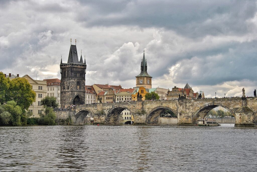 Stone arches of the historic Charles Bridge span the Vltava River in Prague, with the Old Town Bridge Tower and a clock tower rising among colorful buildings under a cloudy sky. Tourists walk across the bridge while a boat passes below.