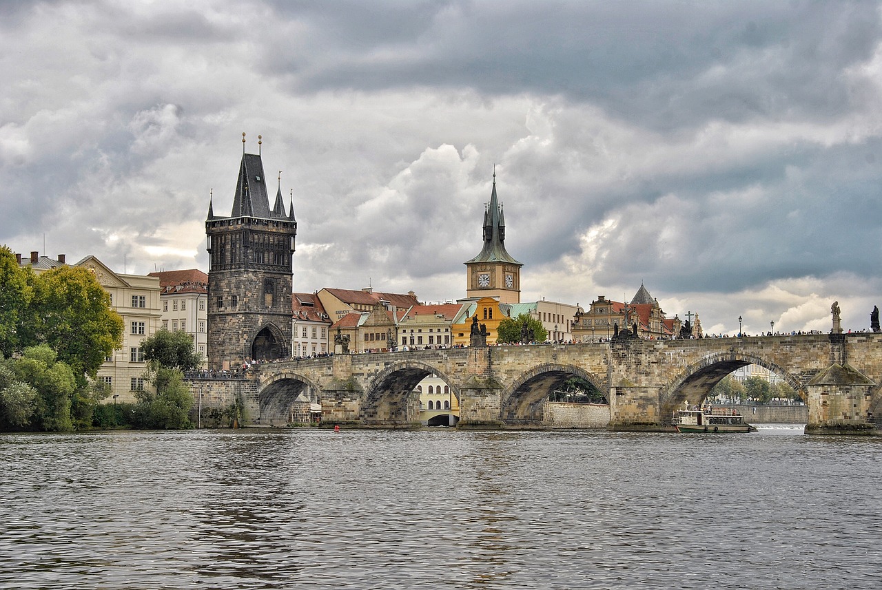 Stone arches of the historic Charles Bridge span the Vltava River in Prague, with the Old Town Bridge Tower and a clock tower rising among colorful buildings under a cloudy sky. Tourists walk across the bridge while a boat passes below.