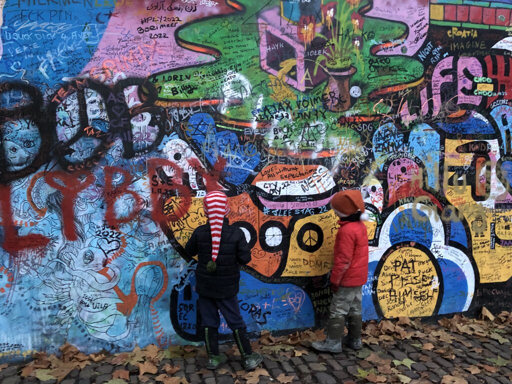 Two children stand in front of the John Lennon Wall in Prague, which is covered in vibrant, overlapping graffiti, tags, peace symbols, and messages in various languages. Fallen autumn leaves line the cobblestone ground beneath them.