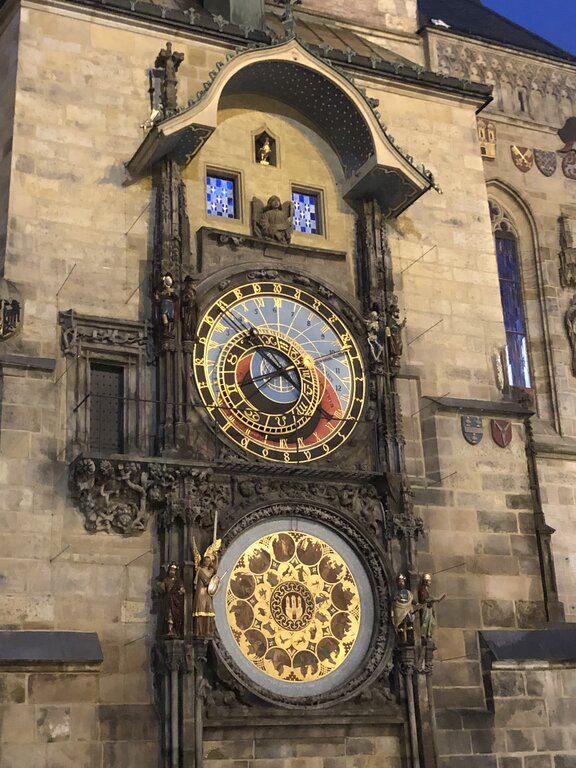 The Prague Astronomical Clock, or Orloj, is illuminated at dusk on the Old Town Hall’s stone facade. The intricate medieval timepiece features a golden astronomical dial with Roman numerals and zodiac signs, along with a lower calendar dial adorned with detailed symbols. Small statues of saints and allegorical figures surround the clock, adding to its Gothic architectural beauty.