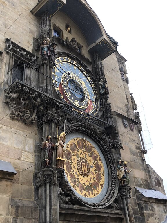 The Prague Astronomical Clock, or Orloj, is mounted on the Old Town Hall in Prague, Czech Republic. The intricate medieval clock features a large blue and gold astronomical dial displaying time, zodiac signs, and planetary positions, with a lower calendar dial adorned with ornate symbols. Surrounding the clock are detailed sculptures, including figures representing virtues and vices, adding to its Gothic charm.