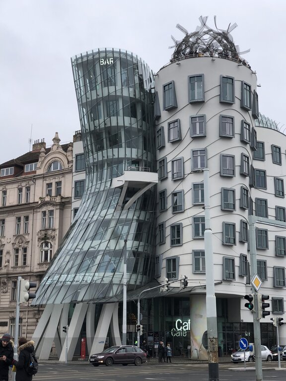 The Dancing House in Prague, an iconic deconstructivist building, features two uniquely shaped towers that appear to lean into each other. One tower, made of curved glass, resembles a woman in motion, while the other has a more rigid, wavy façade with irregularly placed windows and a metal sculpture on top. Designed by architects Frank Gehry and Vlado Milunić, the structure stands in striking contrast to the historic buildings surrounding it.
