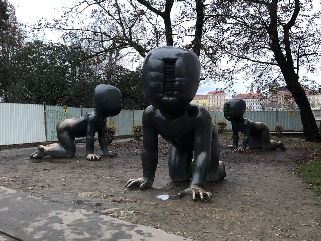 Three large, bronze crawling baby sculptures by artist David Černý are displayed in a park on Prague’s Kampa Island. The figures have exaggerated, featureless heads with deep, vertical slits where their faces should be, giving them an eerie, surreal appearance. The overcast sky and muddy ground add to the striking and unsettling atmosphere of the installation.