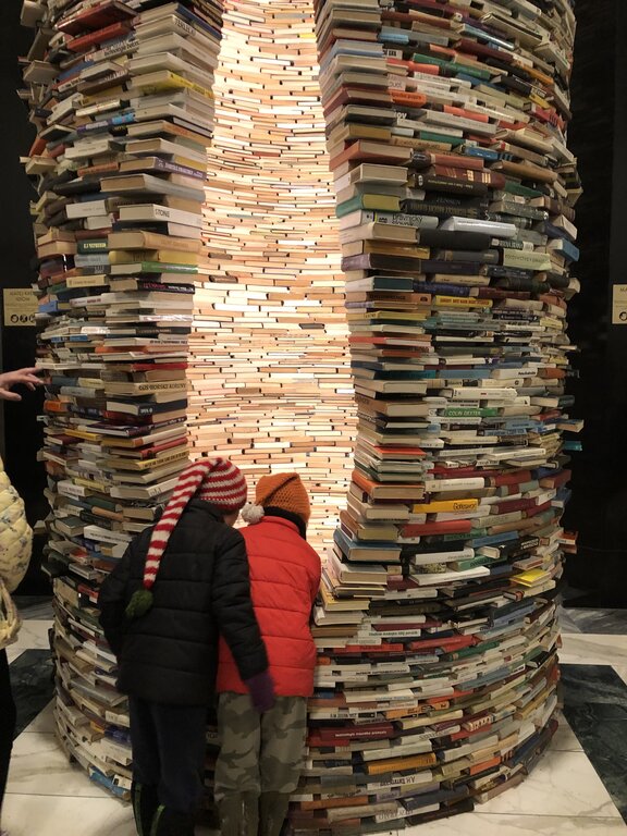 A towering sculpture made of stacked books, known as the Idiom installation, stands in the Prague Municipal Library. The hollow interior, illuminated from within, creates an illusion of endless books through strategically placed mirrors. Two children in colorful winter hats peer into the structure, adding a sense of curiosity and wonder.