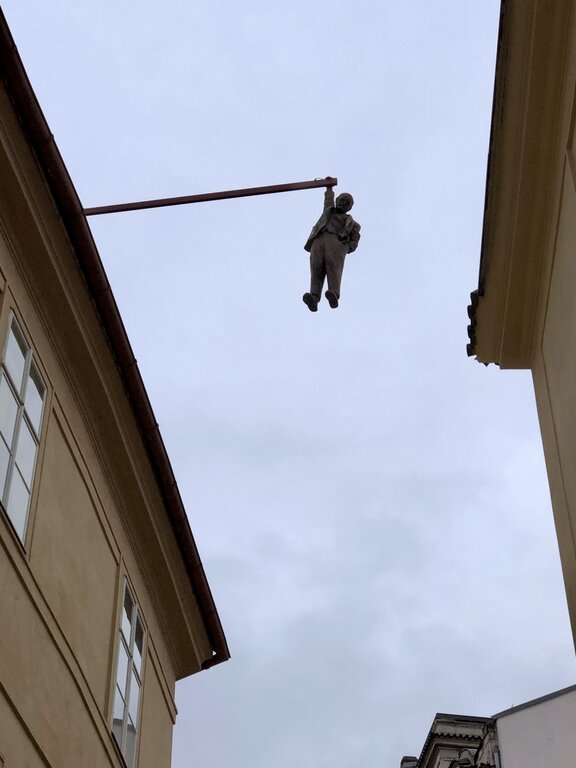 A life-sized sculpture of a man in a suit hangs one-handed from a metal beam extending over a street in Prague. Created by artist David Černý, the artwork, titled Man Hanging Out, depicts Sigmund Freud, symbolizing intellectual struggle and existential uncertainty. The statue appears strikingly realistic against the backdrop of historic buildings and the overcast sky.