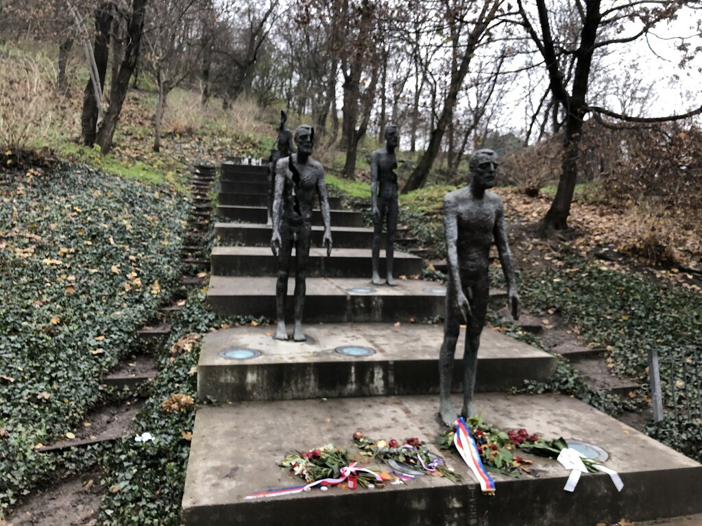 The Memorial to the Victims of Communism in Prague features a series of bronze statues descending a concrete staircase. Each figure appears increasingly fragmented and deteriorated, symbolizing the suffering and loss experienced under communist rule. Flowers and ribbons are placed at the base of the memorial, honoring those who were persecuted.