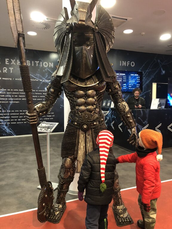 A towering metal sculpture of Anubis, the ancient Egyptian god of the afterlife, stands inside the Museum of Steel Figures in Prague. The intricately detailed figure, made from recycled metal parts, holds a long staff and wears a jackal-headed helmet. Two children in colorful winter hats stand in front of the statue, reaching out in fascination.