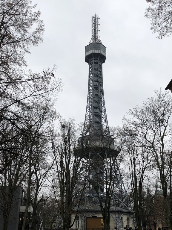 The Petřín Tower in Prague, a steel observation tower resembling a smaller version of the Eiffel Tower, stands tall against an overcast sky. Surrounded by leafless trees, the tower's intricate lattice structure and viewing platform near the top are visible. Built in 1891, this landmark offers panoramic views of the city from its hilltop location.