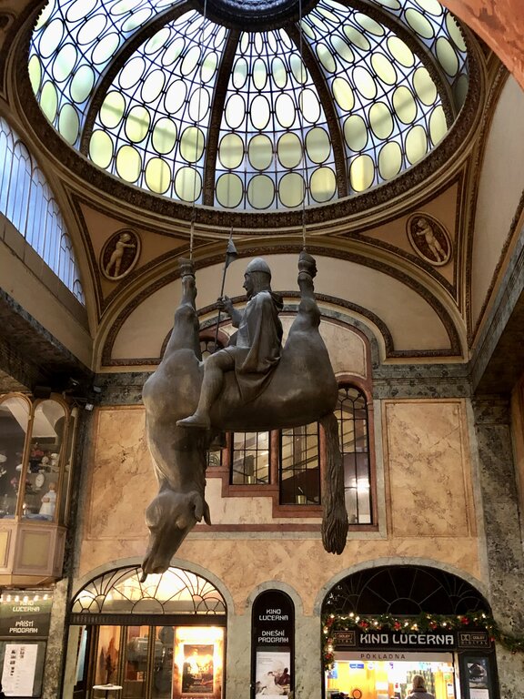 A striking sculpture of Saint Wenceslas riding an upside-down, lifeless horse hangs from the ceiling of Lucerna Passage in Prague, Czech Republic. Created by artist David Černý, this satirical artwork is a playful twist on the traditional equestrian statue, with Wenceslas calmly seated atop the belly of the limp, inverted horse. The elegant glass dome and ornate Art Nouveau architecture of the passage provide a dramatic backdrop to the surreal installation.