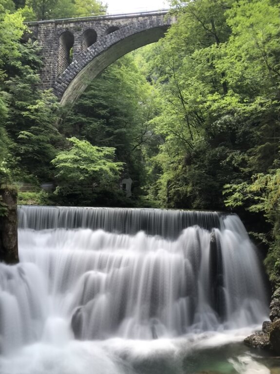 A cascading waterfall flows smoothly over a man-made dam in Vintgar Gorge, Slovenia, surrounded by lush green foliage. Above the waterfall, a historic stone railway bridge with elegant arches spans the gorge, blending harmoniously with the natural scenery. The soft, misty effect of the flowing water contrasts with the rugged stone architecture and dense forest backdrop.