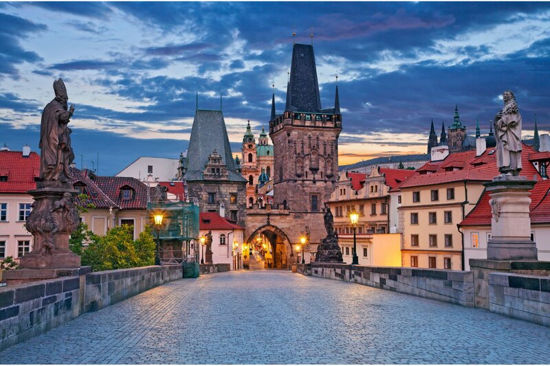 A stunning twilight view of Charles Bridge in Prague, leading towards the Lesser Town (Malá Strana) with its historic buildings and Gothic towers. The cobblestone pathway is lined with Baroque statues, illuminated by glowing street lamps, creating a warm and inviting atmosphere. The sky is painted with deep blue and orange hues, adding to the fairytale charm of this iconic European destination.