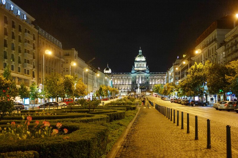 A nighttime view of Wenceslas Square in Prague, with the illuminated National Museum standing majestically at the end of the broad, tree-lined boulevard. The cobblestone pathways are flanked by manicured gardens, street lamps, and historic buildings, while parked cars and pedestrians add to the lively city atmosphere. The glowing lights highlight the grandeur of this famous cultural and commercial hub.