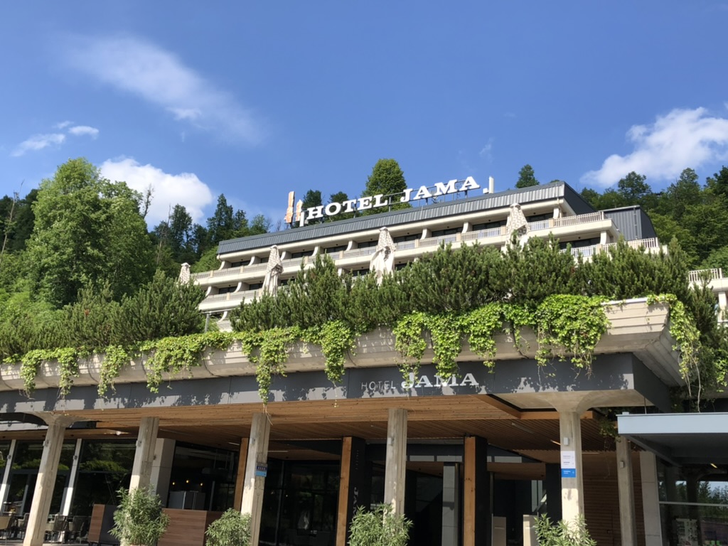 Large hillside hotel with the sign Hotel Jama on the roof, featuring multiple balconies and a modern concrete entrance covered in cascading green vines. The building sits against a backdrop of dense trees and a bright blue sky, highlighting its scenic forest setting and welcoming exterior.