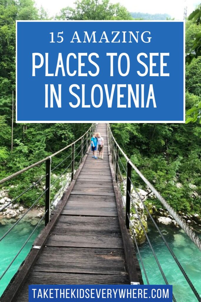Two children stand together on a narrow wooden suspension bridge crossing a bright turquoise river surrounded by dense green forest. Large blue text overlay reads "15 AMAZING PLACES TO SEE IN SLOVENIA" and a blue banner at the bottom reads "TAKETHEKIDSEVERYWHERE.COM".