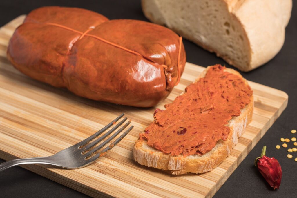 A wooden cutting board holds a spread of spicy 'Nduja, a soft, spreadable Calabrian sausage, generously smeared on a slice of rustic bread. Beside it, the whole sausage is displayed with its signature reddish-orange color. A fork with traces of 'Nduja, a dried chili pepper, and scattered chili seeds hint at the rich, spicy flavors of this traditional Italian delicacy.