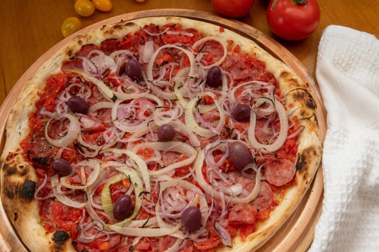 A traditional Calabrian-style pizza with a crispy, charred crust, topped with finely chopped tomatoes, thinly sliced onions, spicy salami, and dark purple olives. The vibrant toppings contrast against the golden-brown dough, creating a rustic and flavorful presentation. Fresh tomatoes and a white kitchen towel rest beside the wooden pizza tray, enhancing the homemade feel.