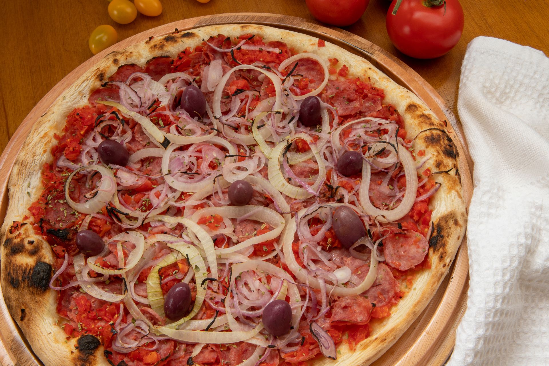 A traditional Calabrian-style pizza with a crispy, charred crust, topped with finely chopped tomatoes, thinly sliced onions, spicy salami, and dark purple olives. The vibrant toppings contrast against the golden-brown dough, creating a rustic and flavorful presentation. Fresh tomatoes and a white kitchen towel rest beside the wooden pizza tray, enhancing the homemade feel.