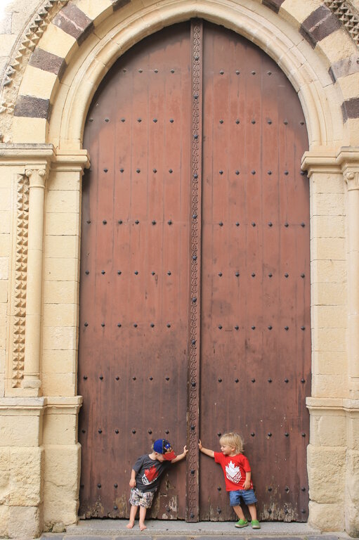 Two young children stand in front of a massive wooden door with intricate iron details, set within a grand arched stone entrance. One child, wearing a red maple leaf shirt, playfully pulls on the door handle, while the other, dressed in a dark maple leaf shirt, leans curiously against the door. The historic architecture, with its ornate stone carvings and aged wood, creates a striking contrast to their small figures.