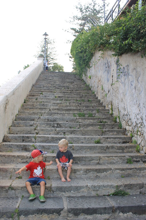 Two young children sit on a long, weathered stone staircase, engaged in conversation. One wears a red cap and matching maple leaf shirt, gesturing animatedly, while the other, in a dark maple leaf shirt, listens intently with bare feet. The old staircase, lined with a white wall and greenery, has patches of grass growing between the cracks, adding to the rustic charm of the scene.