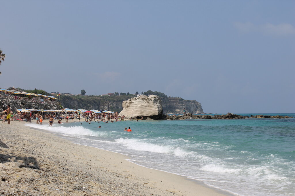 A beautiful beach in Zambrone, Italy, with soft golden sand and turquoise waves gently rolling onto the shore. The beach is filled with sunbathers, colorful umbrellas, and swimmers enjoying the warm Mediterranean waters. In the distance, a striking white rock formation juts out from the sea, with rocky cliffs and lush greenery providing a scenic backdrop.