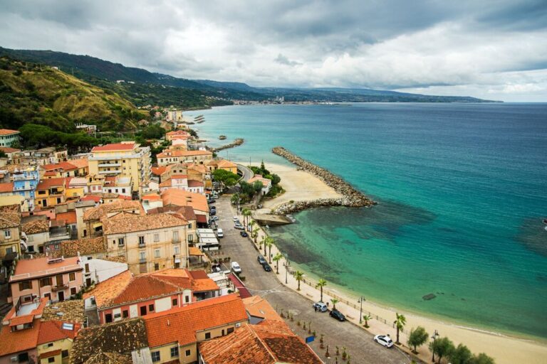 A panoramic view of Pizzo, Italy, showcasing terracotta-roofed buildings cascading down toward a sandy beach and turquoise sea. A stone jetty extends into the water, while the coastline curves toward distant hills under a moody, cloud-filled sky.