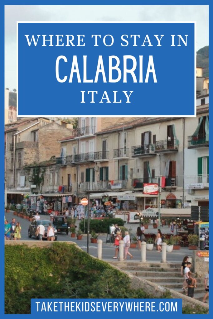 Coastal town in Calabria with pedestrians, shops, balconies, and hillside buildings overlooking a waterfront promenade. The graphic includes the text “WHERE TO STAY IN CALABRIA ITALY” and “takethekidseverywhere.com.”