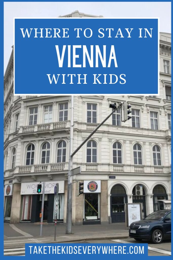 Large white historic building on a city street in Vienna with storefronts, traffic lights, and a parked vehicle in front. The graphic includes the text “WHERE TO STAY IN VIENNA WITH KIDS” and “takethekidseverywhere.com.”
