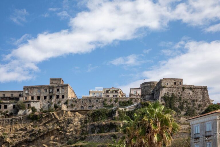 A cluster of historic stone buildings, including a weathered fortress, sits atop a rocky cliff in Pizzo, Italy. The structures are backed by a bright blue sky with scattered clouds, while palm trees and newer homes frame the foreground.