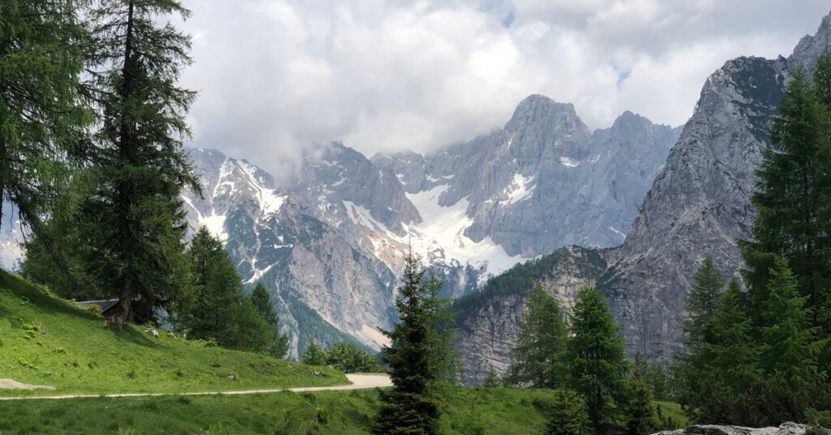 A breathtaking view from the Vršič Pass in Slovenia, showing a winding mountain road cutting through lush green meadows and pine trees, with dramatic, jagged peaks of the Julian Alps in the background partially shrouded in mist and clouds. Snow patches remain nestled in the rocky slopes, adding contrast to the summer landscape.