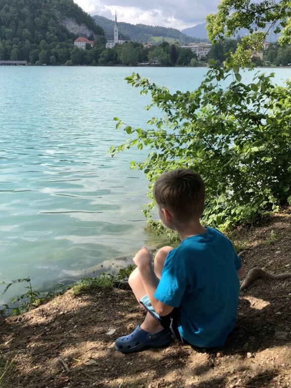A young boy in a blue shirt and Crocs sits quietly at the edge of Lake Bled, gazing across the turquoise water toward a hillside church and village set on the edge of the lake