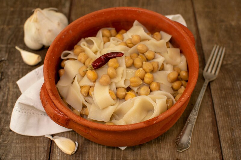 A rustic terracotta bowl filled with lagane e ceci, a traditional Calabria pasta dish made with wide, flat noodles and chickpeas. The dish is garnished with a whole dried chili pepper, and surrounded by garlic cloves and a vintage fork on a wooden table.