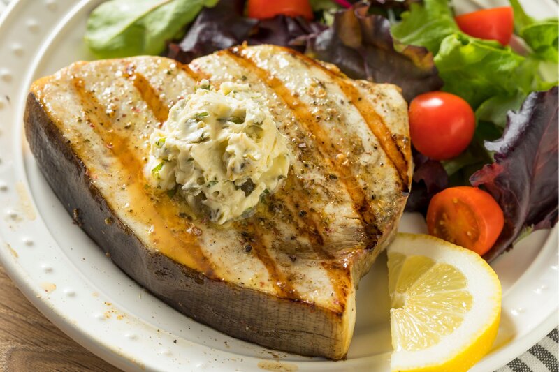 Grilled swordfish steak served with a dollop of herb butter on top, accompanied by a fresh salad of cherry tomatoes and mixed greens. A wedge of lemon sits on the side, adding a bright citrus option to the savoury, lightly charred seafood dish.