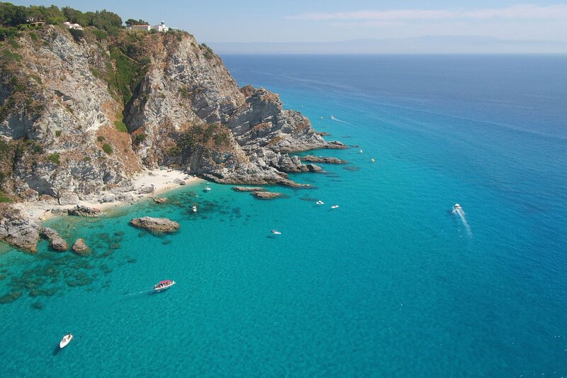 A stunning aerial view of Capo Vaticano's rugged cliffs dropping into vibrant turquoise waters, with white-sand coves tucked between rocky outcrops. Several small boats float in the clear sea, and a white lighthouse sits atop the cliff surrounded by trees.