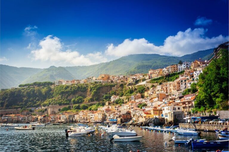 Vibrant hillside homes in Chianalea, a town that's a must see on yourCalabria itinerary, cascade toward the sea, where small boats float peacefully in the harbor. Lush green mountains rise in the background under a bright blue sky dotted with soft white clouds.