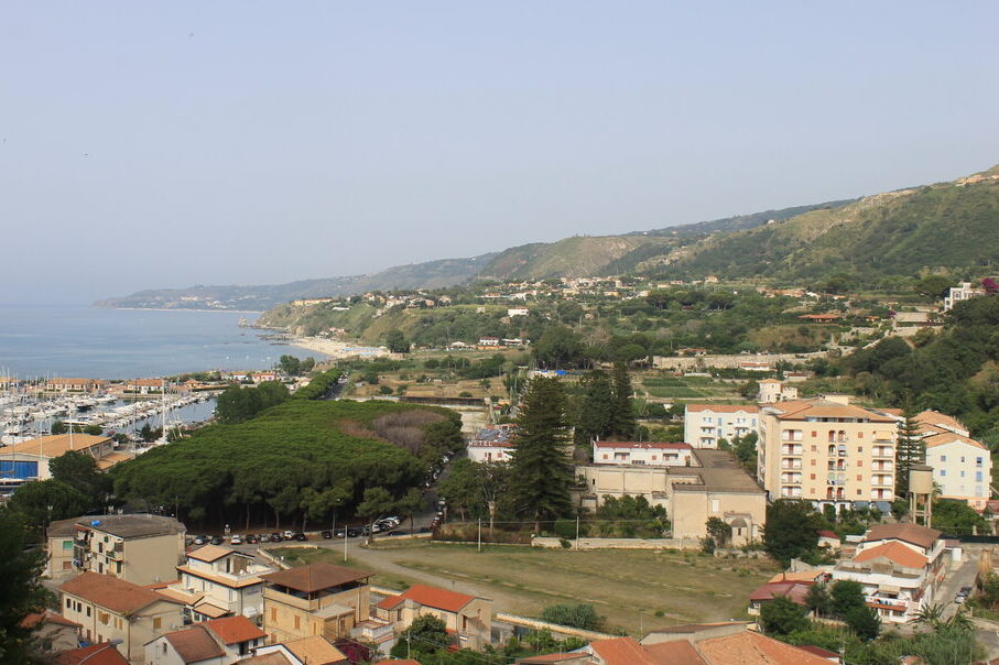 A scenic coastal view of Vibo Marina in Calabria, Italy, featuring a marina filled with boats, clusters of terracotta-roofed buildings, and rolling green hills that stretch along the shoreline.
