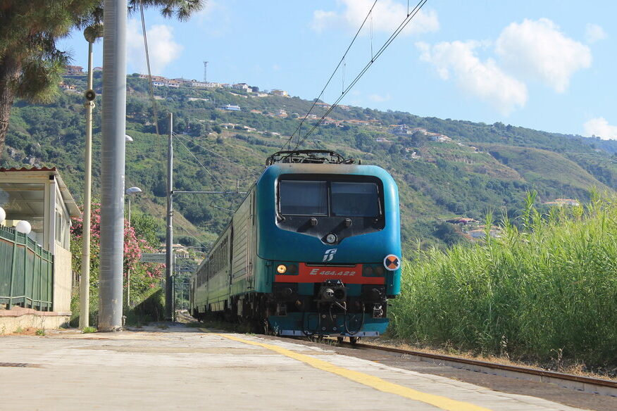 A Trenitalia regional train approaches a small countryside station in Calabria, flanked by tall greenery and backed by terraced hills with scattered houses under a bright blue sky with scattered clouds.
