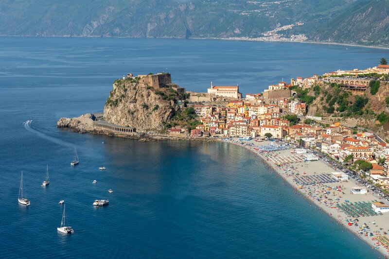 Aerial view of Scilla, a picturesque seaside town in Calabria, Italy, with colorful houses clustered around a dramatic cliff topped by Castello Ruffo. The beach below is lined with umbrellas and sunbathers, while sailboats drift across the crystal-clear blue waters of the Tyrrhenian Sea.