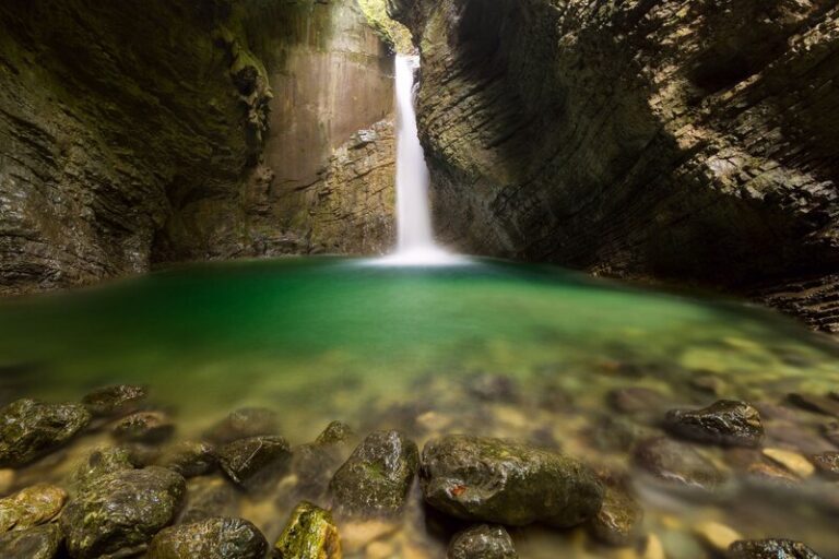 A narrow waterfall plunges into a vibrant emerald-green pool nestled within a steep, rocky gorge in Slovenia. Smooth stones line the water’s edge in the foreground, while the cavern’s textured rock walls curve inward, creating a secluded and tranquil atmosphere.