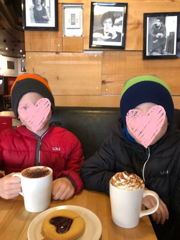 Two children in winter hats and puffy jackets sit at a wooden table inside a cozy café in Quebec City in winter. Each holds a large mug of hot chocolate topped with whipped cream while a donut with chocolate filling sits on a plate between them, with framed black and white portraits hanging on the wooden wall behind them.